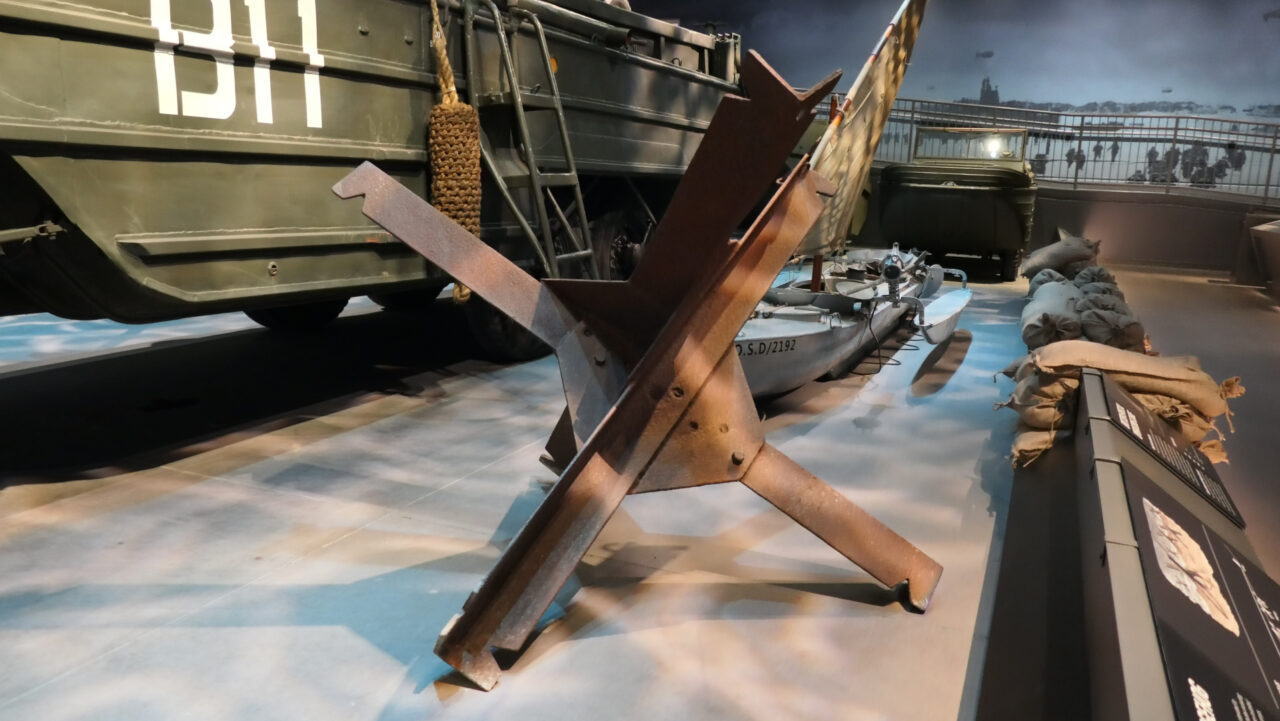 A World War II-era beach landing diorama featuring a Czech hedgehog tank trap in the foreground, positioned in front of a green amphibious landing craft marked "B11." Behind it are military equipment including an inflatable boat, sandbags, and a backdrop mural depicting soldiers storming a beach under aerial support—evoking the D-Day landings of Normandy. Exhibited at the National Museum of Military Vehicles.